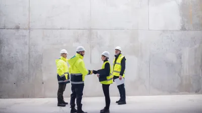 Four construction workers wearing high-visibility jackets and helmets standing and talking near a large concrete wall.