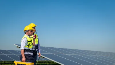 A construction worker wearing a yellow hard hat and safety vest stands beside a solar panel array, holding a toolbox in a sunny outdoor environment.