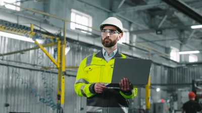 Person in a hard hat and high-visibility jacket using a laptop at an industrial facility.