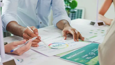 Close-up of hands reviewing architectural or technical documents with charts and drawings on a desk, suggesting a collaborative planning or design meeting.