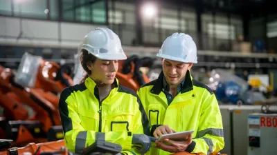 Two workers in high-visibility jackets and hard hats inspecting robotic equipment in a factory.