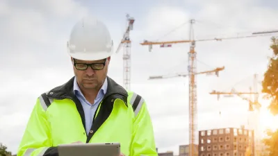 A person in a high-visibility jacket and white hard hat using a tablet on a construction site, with cranes and buildings in the background at sunset.