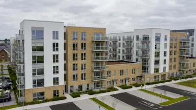 Exterior view of a modern apartment complex with a mix of white and brick facades, multiple balconies with glass railings, and a parking area in front.