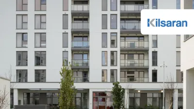Front view of a modern apartment building with several balconies, large windows, and a landscaped entrance area with plants and trees.