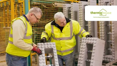 Two people in high-visibility jackets inspecting large grey insulation blocks inside a manufacturing facility, with machinery and safety fencing in the background.