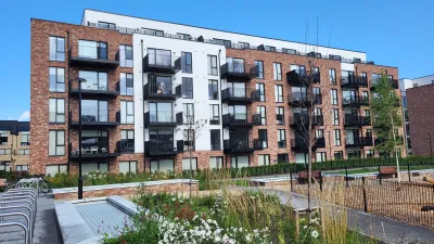 Modern apartment building with a mix of red brick and white facades, featuring multiple black balconies. In the foreground, there is a landscaped garden with flowers, plants, and a pathway under a clear blue sky.