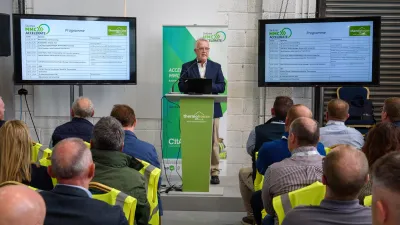 A speaker stands at a podium giving a presentation during an event. Two large screens on either side display the event programme. The audience, many wearing high-visibility vests, is seated and listening.