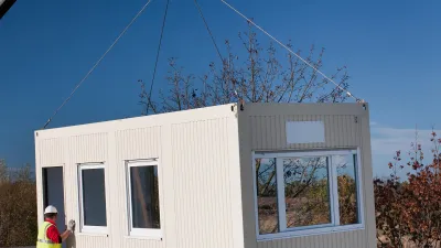 Prefabricated modular unit being lifted by a crane onto a site, with two workers in safety gear guiding it into place.