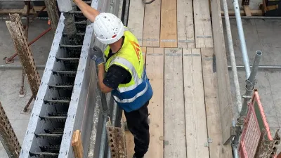 Construction worker in a helmet and high-visibility vest pours concrete into insulated concrete formwork on a building site.