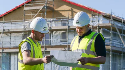 Two construction workers in high-visibility vests and MMC Eco Homes helmets reviewing building plans in front of a house under scaffolding.