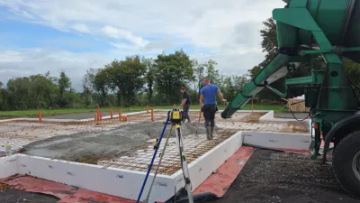 Two people working on site as concrete is poured from a green truck into insulated concrete form foundations with visible rebar.
