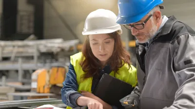 Two people wearing hard hats and safety vests reviewing documents at a worksite, with one person pointing at the papers.