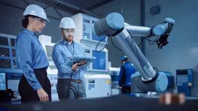Two engineers in hard hats use a tablet to program a robotic arm on a factory floor.