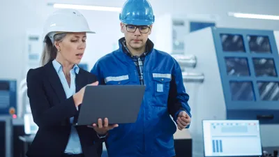 Two people wearing hard hats in an industrial setting, one in a business suit holding a laptop and the other in a blue work jacket, reviewing information together.
