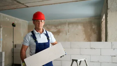 Construction worker in a red hard hat and overalls holding a large white block indoors, with a step ladder and unfinished wall in the background.