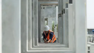"Person wearing a hard hat and orange safety vest working with a measuring tool inside a series of concrete rectangular frames at a construction site.