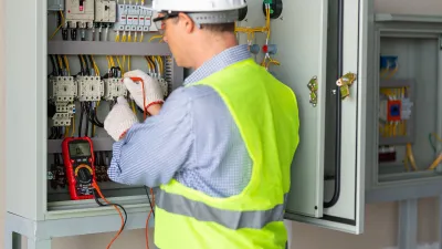 Person wearing a hard hat, high-visibility vest, and gloves using a multimeter to check electrical connections inside an open control panel.