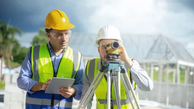 Two people wearing hard hats and reflective vests working at a construction site, with one person using a surveyor’s level on a tripod and the other holding a tablet.