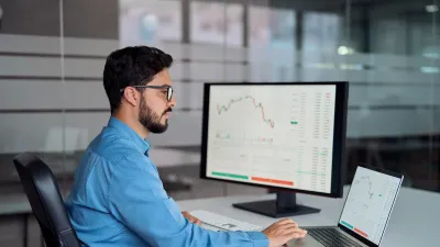 Person in a blue shirt working at a desk with a laptop and large monitor displaying financial charts and data.