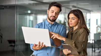 Two people standing in an office, holding a laptop and a tablet, discussing something on the laptop screen.