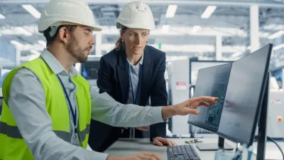 Engineer in a safety vest and hard hat pointing at a computer screen while discussing data with a colleague in a hard hat inside an industrial facility.