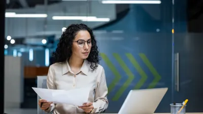 Person with curly hair and glasses sitting at a desk, holding papers and looking at a laptop screen in an office setting.