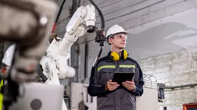 Person wearing a hard hat, safety glasses, and protective clothing with yellow ear protectors around their neck, holding a tablet while standing next to industrial robotic machinery.