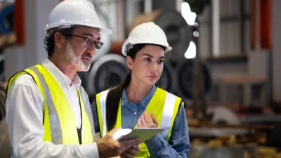 Two people wearing white hard hats and yellow reflective vests standing in an industrial setting, looking in the same direction while one holds a tablet.