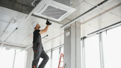 Person wearing a hard hat and overalls standing on a ladder and installing or servicing a ceiling-mounted air conditioning unit in a modern building.