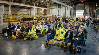 A large group of workers in safety vests gathered inside an industrial manufacturing facility.
