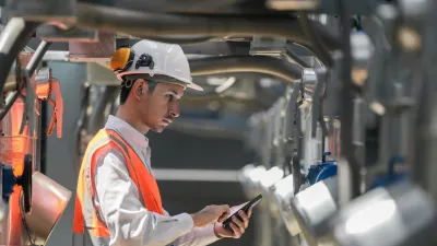 A construction worker wearing safety gear using a tablet near machinery.