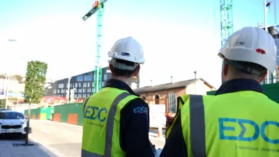 Two people wearing hard hats and high-visibility vests observing a construction site with cranes and modern buildings.