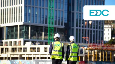 Two people wearing hard hats and high-visibility vests with the EDC logo, observing a modern glass building under construction.