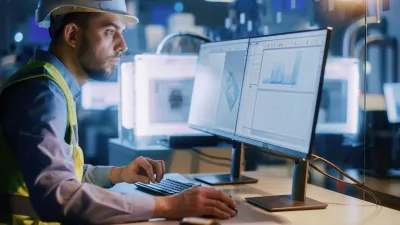 Construction worker with hard hat using multiple computer monitors in a dark room.