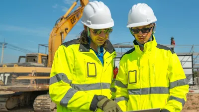 Two construction workers in hard hats and high-vis jackets reviewing plans at a building site.