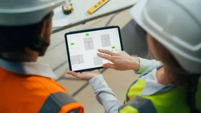 Two construction workers in safety helmets reviewing plans on a tablet at a worksite.