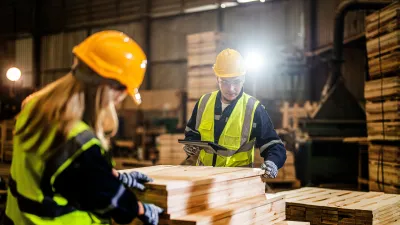 Worker in yellow hard hat and safety vest examining wooden planks in a warehouse setting.
