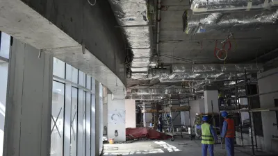 Construction site interior showing exposed ceiling with pipes and ductwork, and unfinished concrete walls.