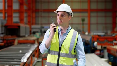 Worker in hard hat and safety vest standing in an industrial warehouse environment.