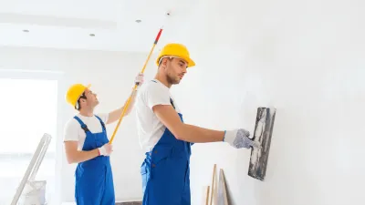 Two workers in yellow hard hats and blue overalls plastering a white interior wall.