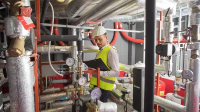 Construction worker in safety vest and helmet working on pipes and valves in industrial setting.
