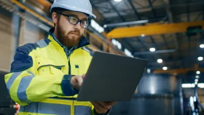 Construction worker in hard hat and safety vest working on laptop inside industrial facility.