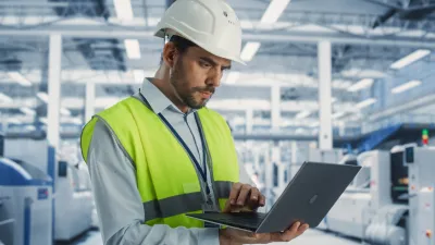 Worker in hard hat and safety vest using laptop in industrial facility.