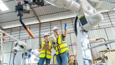 Factory workers in yellow safety vests and hardhats examining industrial robotic equipment in a warehouse.