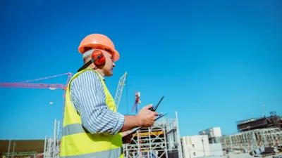 Construction worker in safety vest and helmet using tablet at building site against blue sky.