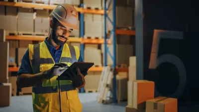Warehouse worker in yellow safety vest using digital tablet among shelving units.