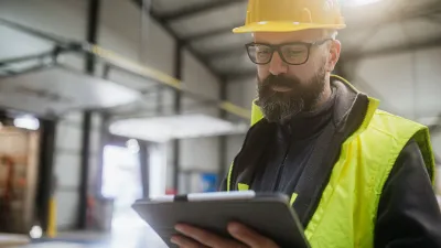 Construction worker in yellow hardhat and safety vest reviewing information on a tablet.