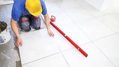 Construction worker in yellow hard hat laying white tile flooring with a red leveling tool.