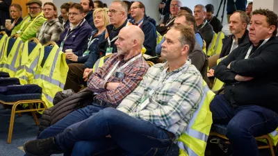 A group of people wearing high-visibility vests seated in an audience at a professional event.