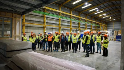 Group of workers in safety vests and helmets standing in a large industrial warehouse with stacked materials.